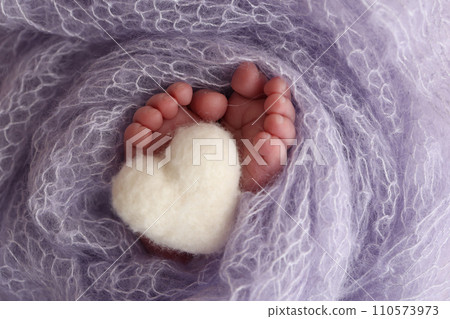 The tiny foot of a newborn baby. Soft feet of a new born in a lilac, purple wool blanket. Close up of toes, heels and feet of a newborn. Knitted white heart in the legs of a baby. Macro photography. The tiny foot of a newborn baby. Soft feet of a new born in a lilac, purple wool blanket. Close up of toes, heels and feet of a newborn. Knitted white heart in the legs of a baby. Macro photography. 110573973