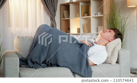 Fever disease. Sickness coughing. High temperature. Ill man wrapped in plaid feeling bad suffering from shivering laying on sofa in living room interior at home. Fever disease. Sickness coughing. High temperature. Ill man wrapped in plaid feeling bad suffering from shivering laying on sofa in living room interior at home. 110574641