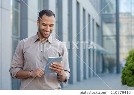 Smiling young man using tablet in urban setting, portraying connectivity, technology, and casual business outdoors. Smiling young man using tablet in urban setting, portraying connectivity, technology, and casual business outdoors. 110579413