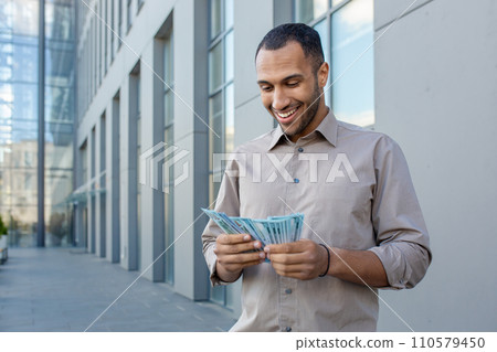 Smiling man counting money outdoors near modern office building, expressing financial success and prosperity Smiling man counting money outdoors near modern office building, expressing financial success and prosperity 110579450