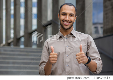 Confident young man in a casual shirt giving thumbs up with a smile, outdoor steps in the background, expressing success and positivity. Confident young man in a casual shirt giving thumbs up with a smile, outdoor steps in the background, expressing success and positivity. 110579613