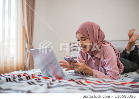 attractive hijab woman using cell phone and laptop while lying on bed attractive hijab woman using cell phone and laptop while lying on bed 110580386