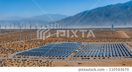 Renewable energy landscape with large wind turbines and solar panels in a sunny, arid desert valley, surrounded by mountains, under a clear blue sky. 110583670