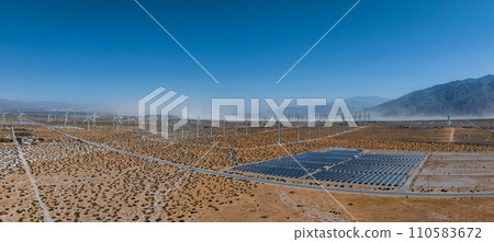 Aerial view of a renewable energy landscape with over 50 wind turbines amidst mountains, under a clear blue sky, with a distant dust storm and desert shrubs. 110583672