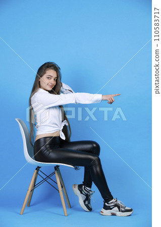 Smiling girl in shirt, pointing away, while looking at camera. Side view of graceful long haired woman gesturing, straightening arm, while resting in chair, isolated on blue. Concept of gesturing. Smiling girl in shirt, pointing away, while looking at camera. Side view of graceful long haired woman gesturing, straightening arm, while resting in chair, isolated on blue. Concept of gesturing. 110583717