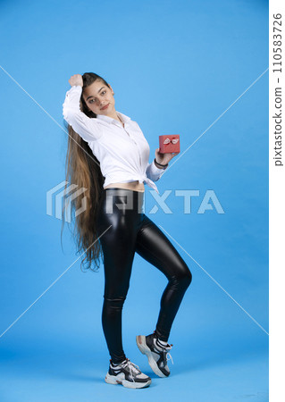 Attractive young woman touching long loose hair, while posing in studio. Front view of graceful birthday girl with small gift box, standing with bent at knee leg, isolated on blue. Concept of posing. Attractive young woman touching long loose hair, while posing in studio. Front view of graceful birthday girl with small gift box, standing with bent at knee leg, isolated on blue. Concept of posing. 110583726