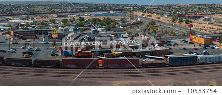 Aerial view of Barstow, California, showcasing Barstow Station, businesses, and vehicles amidst a sunny, semi-arid landscape with hills, clear skies, and scattered greenery. 110583754