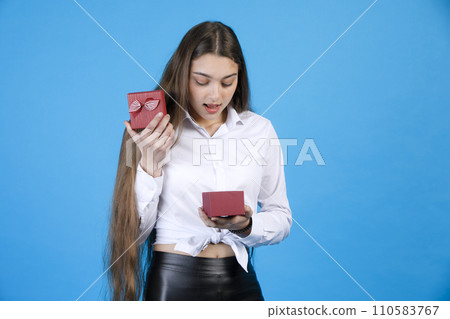 Curious female with long hair opening small red present box, while standing indoors. Surprised happy girl looking into wee gift box, decorated with striped bow, isolated on blue. Concept of present. 110583767