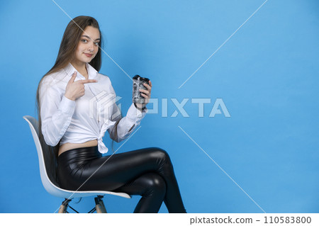 Smiling charming girl in black leggings, pointing at cup of tasty coffee, while sitting in chair. Side view of graceful lady gesturing, while posing, isolated on blue background. Concept of gesturing. Smiling charming girl in black leggings, pointing at cup of tasty coffee, while sitting in chair. Side view of graceful lady gesturing, while posing, isolated on blue background. Concept of gesturing. 110583800