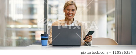 Cheerful female manager use phone while sitting in modern coworking and working on laptop 110586334