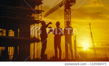 A construction team discussing blueprints at a construction site at sunrise, silhouetted against the dawn sky. 110586473