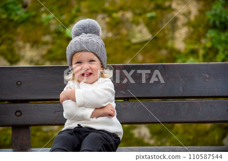 Cute blonde kid sitting on a bench outside in a park, smiling. Happy child wearing a hat outdoors on a spring day. Adorable grinning caucasian boy resting on a bench after play time on a playground. 110587544