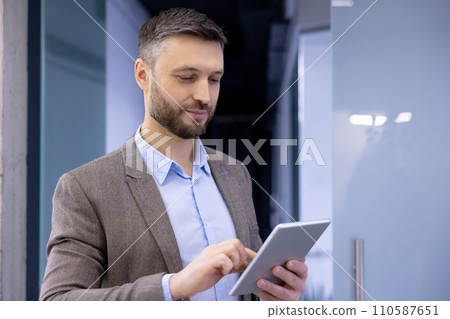 Professional man in smart casual outfit smiling while using a tablet in a modern office setting, depicting work and technology. 110587651