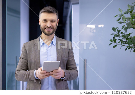 Confident businessman in smart casual attire smiling, holding a tablet in modern office setting, embodying success and professionalism. 110587652