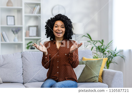 Happy young African American woman sitting on sofa at home, looking, talking and gesturing at camera. 110587721