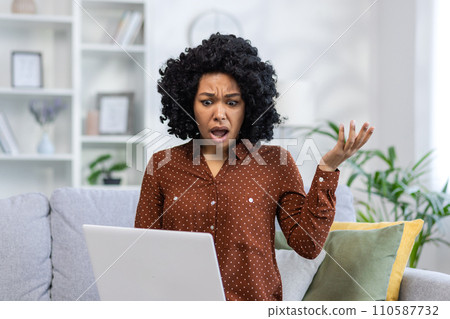 Angry young African American woman sitting on the couch at home and looking at the laptop screen with her hands in frustration. Angry young African American woman sitting on the couch at home and looking at the laptop screen with her hands in frustration. 110587732
