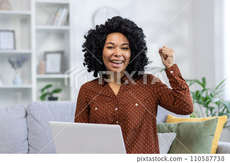 Portrait of a happy African American woman sitting at home on the couch in front of the camera and celebrating her success by raising her hand up 110587738