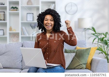 Portrait of a young happy African American woman sitting on the sofa at home, holding a laptop on her lap and looking happy at the camera showing a victory gesture with her hand. Portrait of a young happy African American woman sitting on the sofa at home, holding a laptop on her lap and looking happy at the camera showing a victory gesture with her hand. 110587741