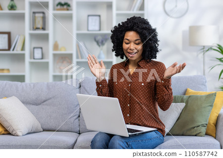 African American young woman sitting on sofa at home and talking smiling and emotional on video call online. African American young woman sitting on sofa at home and talking smiling and emotional on video call online. 110587742