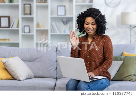 Smiling African American young woman sitting on sofa at home, talking on video call via laptop, waving and greeting at camera. Smiling African American young woman sitting on sofa at home, talking on video call via laptop, waving and greeting at camera. 110587743