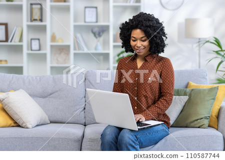 African American young smiling woman sitting on sofa at home, holding laptop, working and studying remotely. 110587744