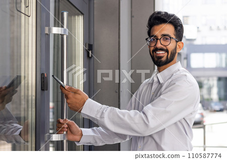 Confident young professional opening office door with smartphone, showcasing modern access technology and a welcoming smile. 110587774