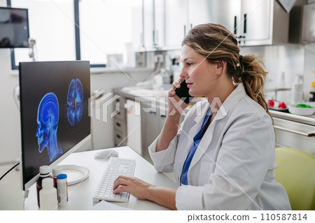 Female doctor working on computer in doctor's office, looking at MRI scan, phone calling test results to patient. Doctor consulting scan with other doctors. 110587814