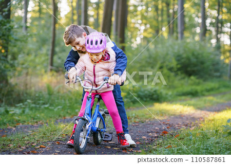 Cute little preschool girl in safety helmet riding bicycle. School kid boy, brother teaching happy healthy sister child cycling and having fun with learning bike. Active siblings family outdoors. Cute little preschool girl in safety helmet riding bicycle. School kid boy, brother teaching happy healthy sister child cycling and having fun with learning bike. Active siblings family outdoors. 110587861