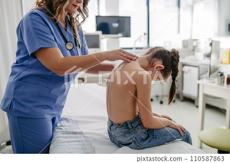Orthopedist examines the spine, posture, and spinal deformities of little girl. Girl visiting paediatrician for annual preventive exam. Concept of preventive health care for children. 110587863