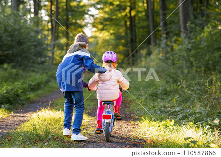 Cute little preschool girl in safety helmet riding bicycle. School kid boy, brother teaching happy healthy sister child cycling and having fun with learning bike. Active siblings family outdoors. 110587866