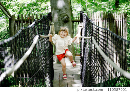 Cute little preschool girl walking on high tree-canopy trail with wooden walkway and ropeways. Happy active child exploring treetop path. Funny activity for families outdoors 110587901