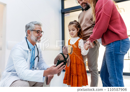 Pediatrician looking at girl's toy. Girl patient is at examination in a modern clinic with her parents. Emotional support during medical examination. Pediatrician looking at girl's toy. Girl patient is at examination in a modern clinic with her parents. Emotional support during medical examination. 110587906