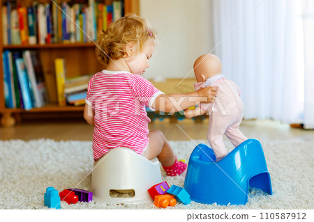 Closeup of cute little 12 months old toddler baby girl child sitting on potty. Kid playing with doll toy. Toilet training concept. Baby learning, development steps Closeup of cute little 12 months old toddler baby girl child sitting on potty. Kid playing with doll toy. Toilet training concept. Baby learning, development steps 110587912