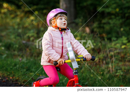 Child riding balance bike. Kids on bicycle in sunny forest. Little girl enjoying to ride glider bike on warm day. Preschooler learning to balance on run bicycle in safe helmet. Sport activity Child riding balance bike. Kids on bicycle in sunny forest. Little girl enjoying to ride glider bike on warm day. Preschooler learning to balance on run bicycle in safe helmet. Sport activity 110587947