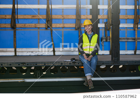 Female warehouse receiver in reflective vest and helmet sitting inside of truck in cargo area, trailer. Female warehouse receiver in reflective vest and helmet sitting inside of truck in cargo area, trailer. 110587962