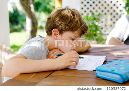 Hard-working sad school kid boy making homework during quarantine time from corona pandemic disease. Upset tired child on home schooling in coronavirus covid time, schools closed. Dad on background Hard-working sad school kid boy making homework during quarantine time from corona pandemic disease. Upset tired child on home schooling in coronavirus covid time, schools closed. Dad on background 110587972