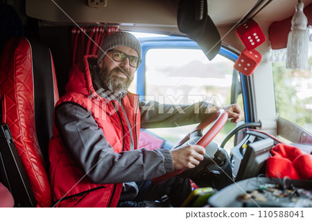 Portrait of confident professional truck driver sitting in truck cabin, looking at camera and smiling. Inside of vehicle. 110588041