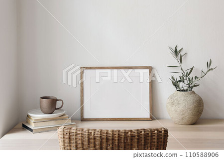Empty horizontal picture frame mockup. Wooden table, desk with cup of coffee, blurred rattan chair. Vase with olive tree branches, old books. Mediterranean interior, home. White wall background. Empty horizontal picture frame mockup. Wooden table, desk with cup of coffee, blurred rattan chair. Vase with olive tree branches, old books. Mediterranean interior, home. White wall background. 110588906
