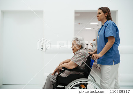 Portrait of nurse pushing senior patient in wheelchair across hospital corridor. Emotional support for elderly woman. 110589517