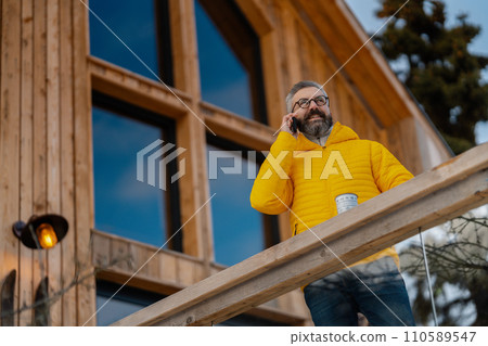 Man phone calling from patio, drinking hot tea, coffee and enjoying beautiful winter day. Handsome man spending relaxing, stress-free winter weekend in cabin in mountains, enjoying alone time. 110589547