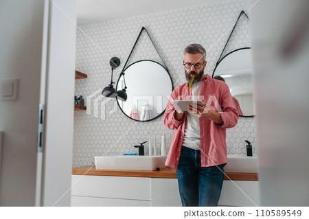 Man shopping online while brushing his teeth in bathroom. Holding tablet, scrolling and buying in eshop, toothbrush in mouth. Man shopping online while brushing his teeth in bathroom. Holding tablet, scrolling and buying in eshop, toothbrush in mouth. 110589549