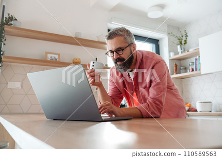 Mature man working from home on laptop, standing by kitchen island, with cup of coffee, holding smartphone. Concept of remote work from home. Hygge at work. Mature man working from home on laptop, standing by kitchen island, with cup of coffee, holding smartphone. Concept of remote work from home. Hygge at work. 110589563