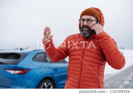 Angry man standing by electric car, battery run out of power before reaching destination. Man phone calling for help waiting for breakdown service car, tow truck. 110589568
