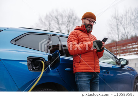 Man charging electric car during cold snowy day, using electric vehicle charging app, checking battery life, energy consumption on smart phone. Charging and driving electric vehicles during winter Man charging electric car during cold snowy day, using electric vehicle charging app, checking battery life, energy consumption on smart phone. Charging and driving electric vehicles during winter 110589593