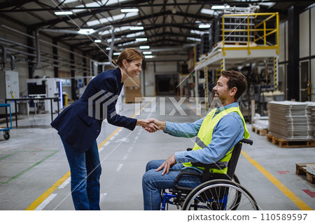 Portrait of man in wheelchair working in warehouse, shaking hand with HR manager, director. Concept of workers with disabilities, accessible workplace for employees with mobility impairment. Portrait of man in wheelchair working in warehouse, shaking hand with HR manager, director. Concept of workers with disabilities, accessible workplace for employees with mobility impairment. 110589597