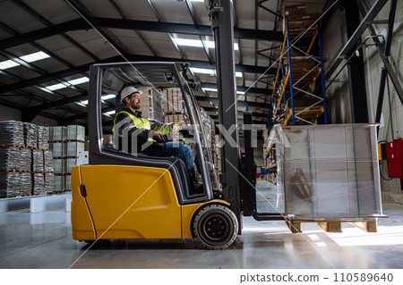 Side view of forklift in warehouse with male driver. Warehouse worker preparing products for shipmennt, delivery, checking stock in warehouse. Side view of forklift in warehouse with male driver. Warehouse worker preparing products for shipmennt, delivery, checking stock in warehouse. 110589640