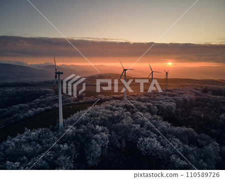 Aerial landscape photography of sunrise over frost-covered nature with wind turbines. Windmill in soft morning light with icy trees around. Concept of wind power as clean, renewable energy source. Aerial landscape photography of sunrise over frost-covered nature with wind turbines. Windmill in soft morning light with icy trees around. Concept of wind power as clean, renewable energy source. 110589726