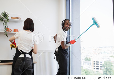 African washing windows with mop, listening to music in headphones and dancing. African washing windows with mop, listening to music in headphones and dancing. 110591356