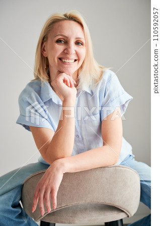 Pretty female relaxing on bar stool. Portrait of woman with blonde hair smiling straight to camera. Beautiful woman sitting on bar stool in her apartment. 110592057