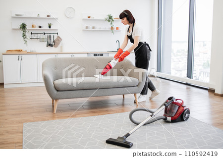 Full length of young cleaner vacuuming sofa with portable vacuum cleaner. 110592419